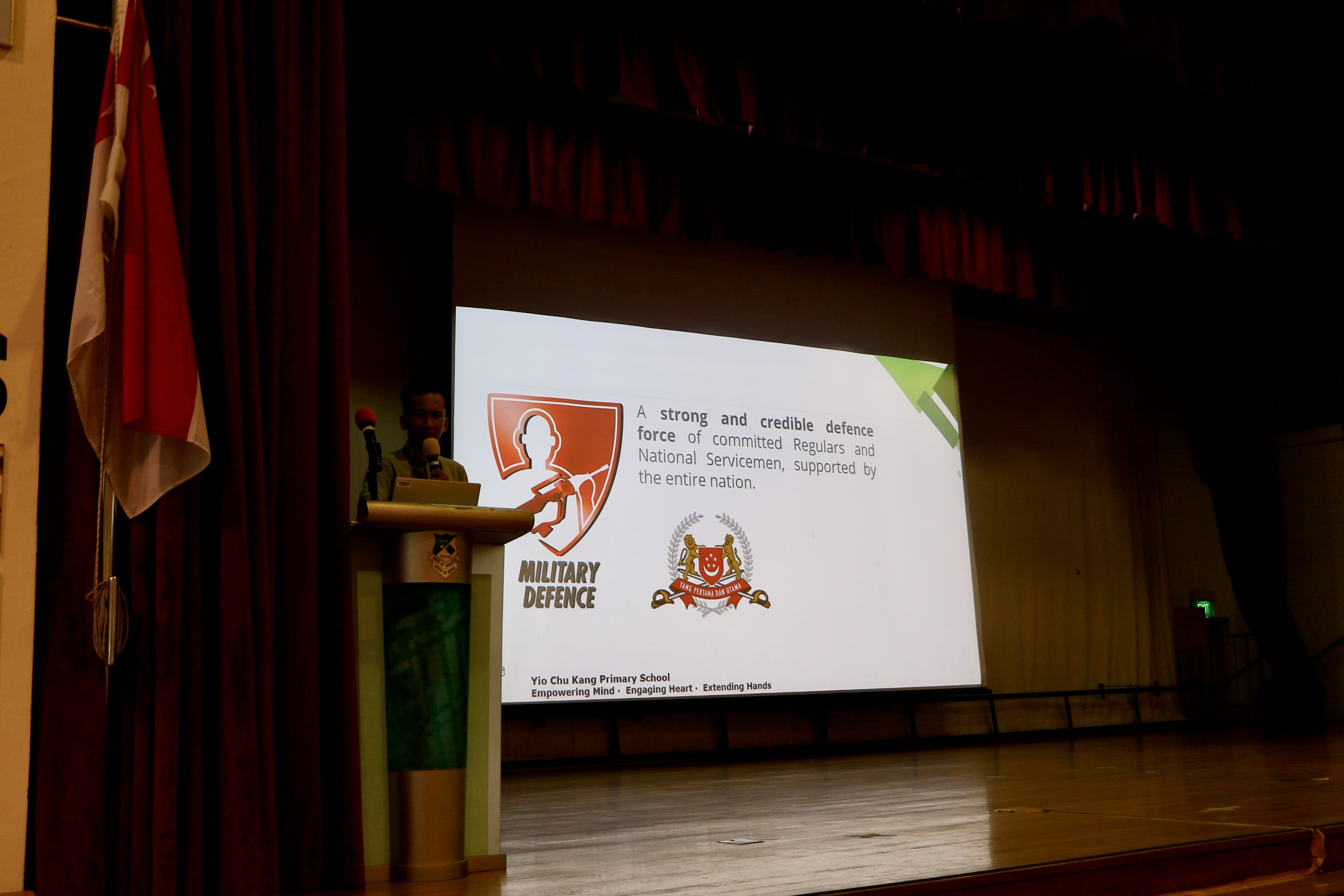 A speaker at a podium on stage presents a Military Defence slideshow at Yio Chu Kang Primary School.